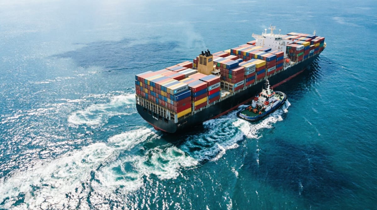An aerial view of a loaded container ship under way at sea, with a tug alongside.